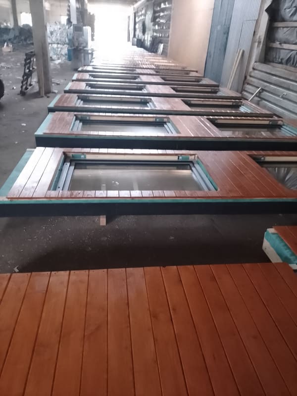 A row of finished wall panels lying flat in the factory hall, each clad in stained wood with a black-framed window already glazed
