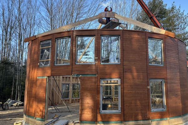 The wall ring standing complete on the crawlspace, a telehandler boom lowering a long timber onto the top of the ring, bare trees and blue sky
