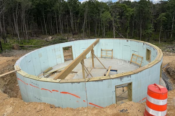 The round basement wall in insulated concrete block, with door and window openings cut, a timber plate on top and bracing across the middle, in cleared woods