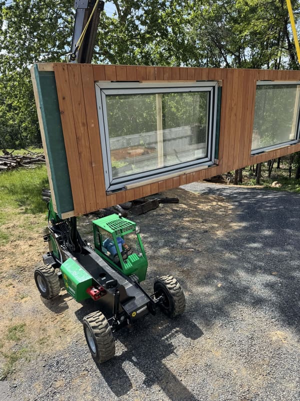 A telehandler carrying one long wood-clad wall panel with two large windows already fitted, on a gravel drive