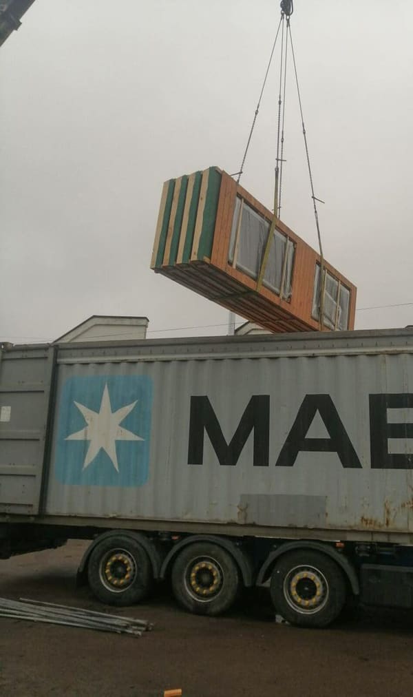 A crane lifting a crated glazed wall panel over a grey 40 ft container on a trailer